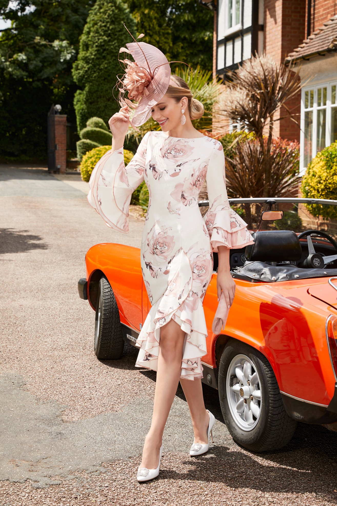 Blonde lady standing in front of red vintage car wearing pink and ivory floral occasion dress with ruffle sleeves and high neckline and matching white high heeled shoes and clutch bag